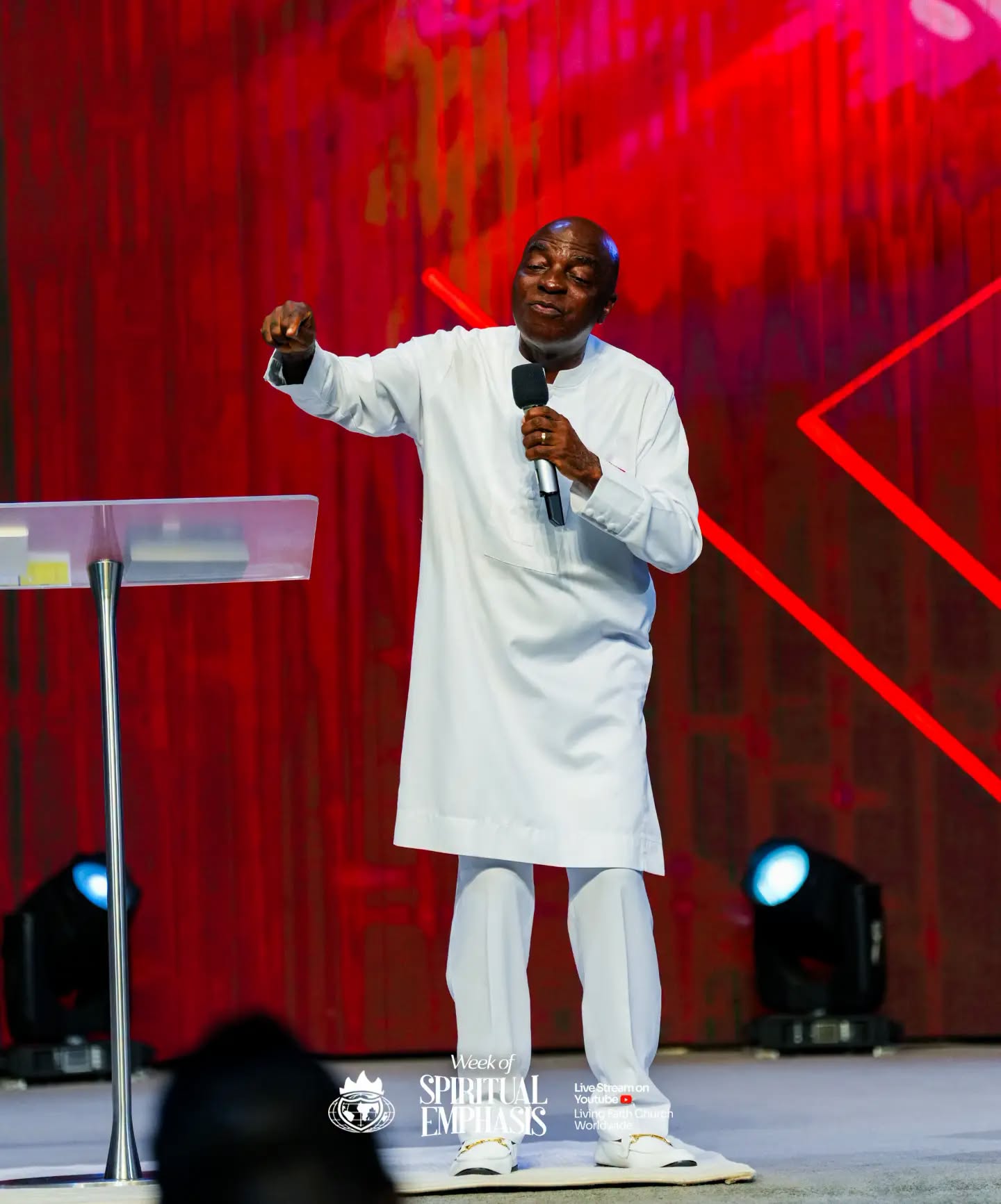 Bishop David Oyedepo preaching into a microphone against a red stage backdrop