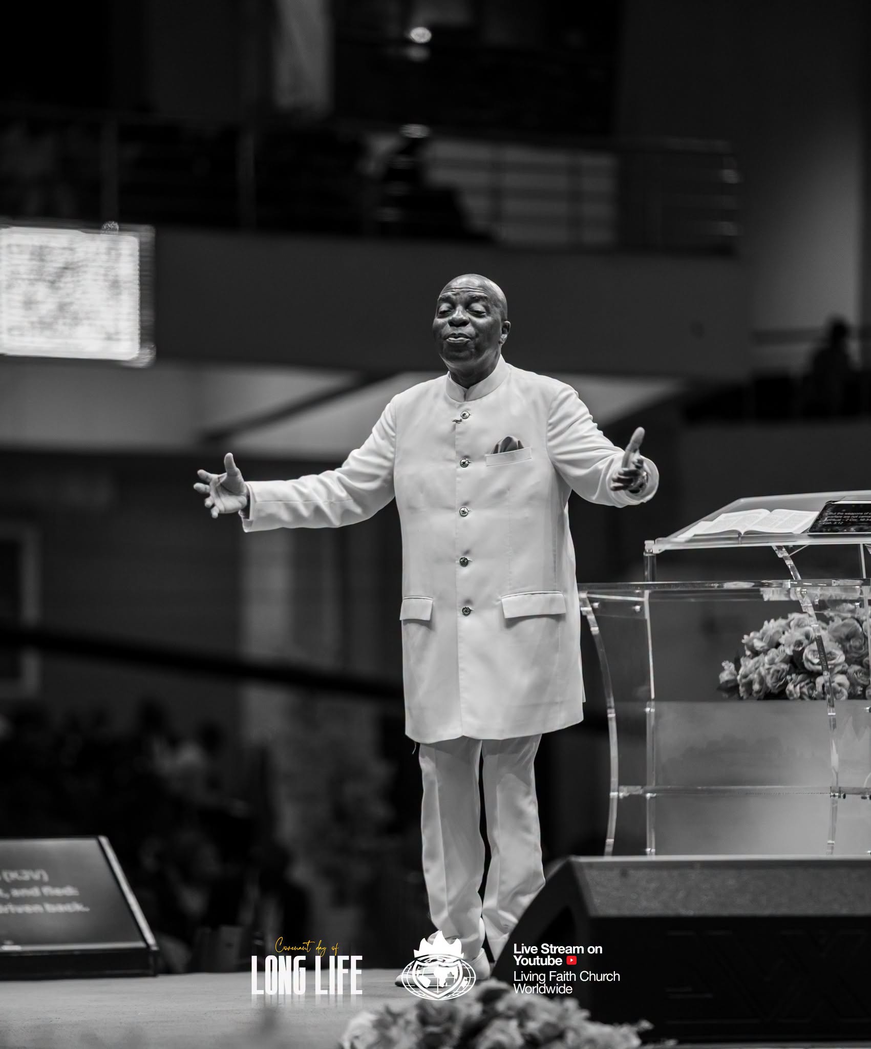 Bishop David Oyedepo with arms open beside the pulpit, in black and white